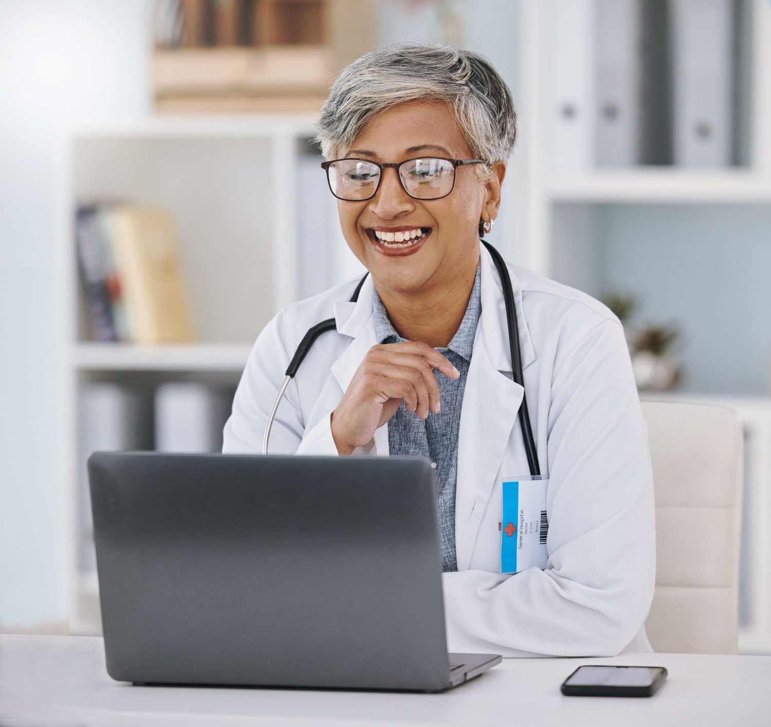 Senior Asian female doctor sitting at her desk on a telehealth visit on her laptop smiling