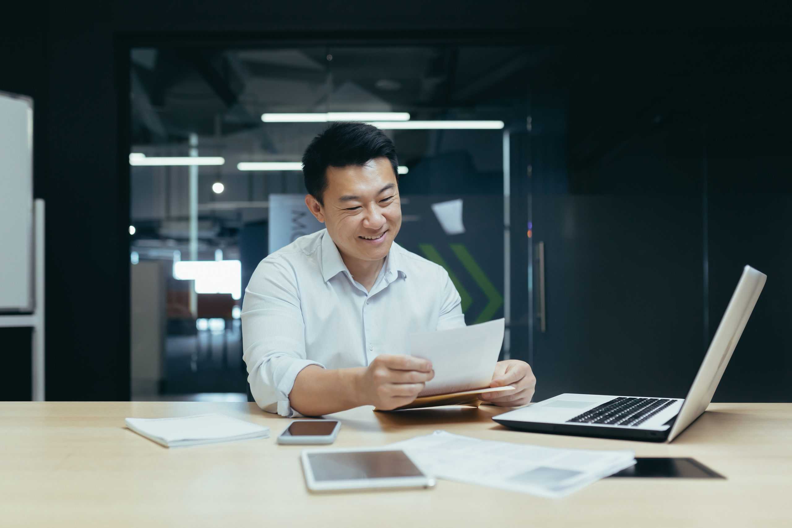 a male professional smiling and reviewing vocational information on paper at his desk with phone and laptop