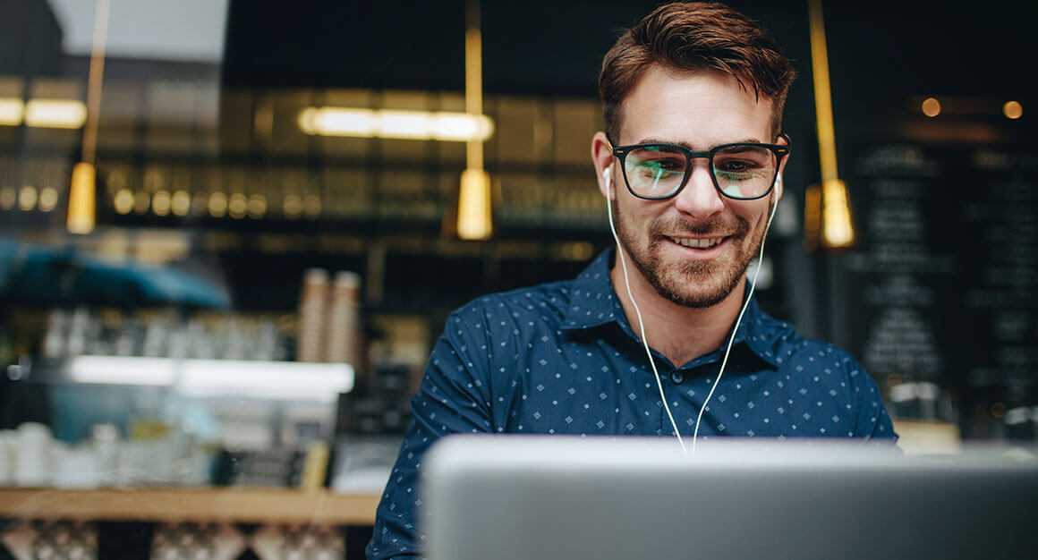 A man with headphone and glasses on, smiling at something on his computer screen