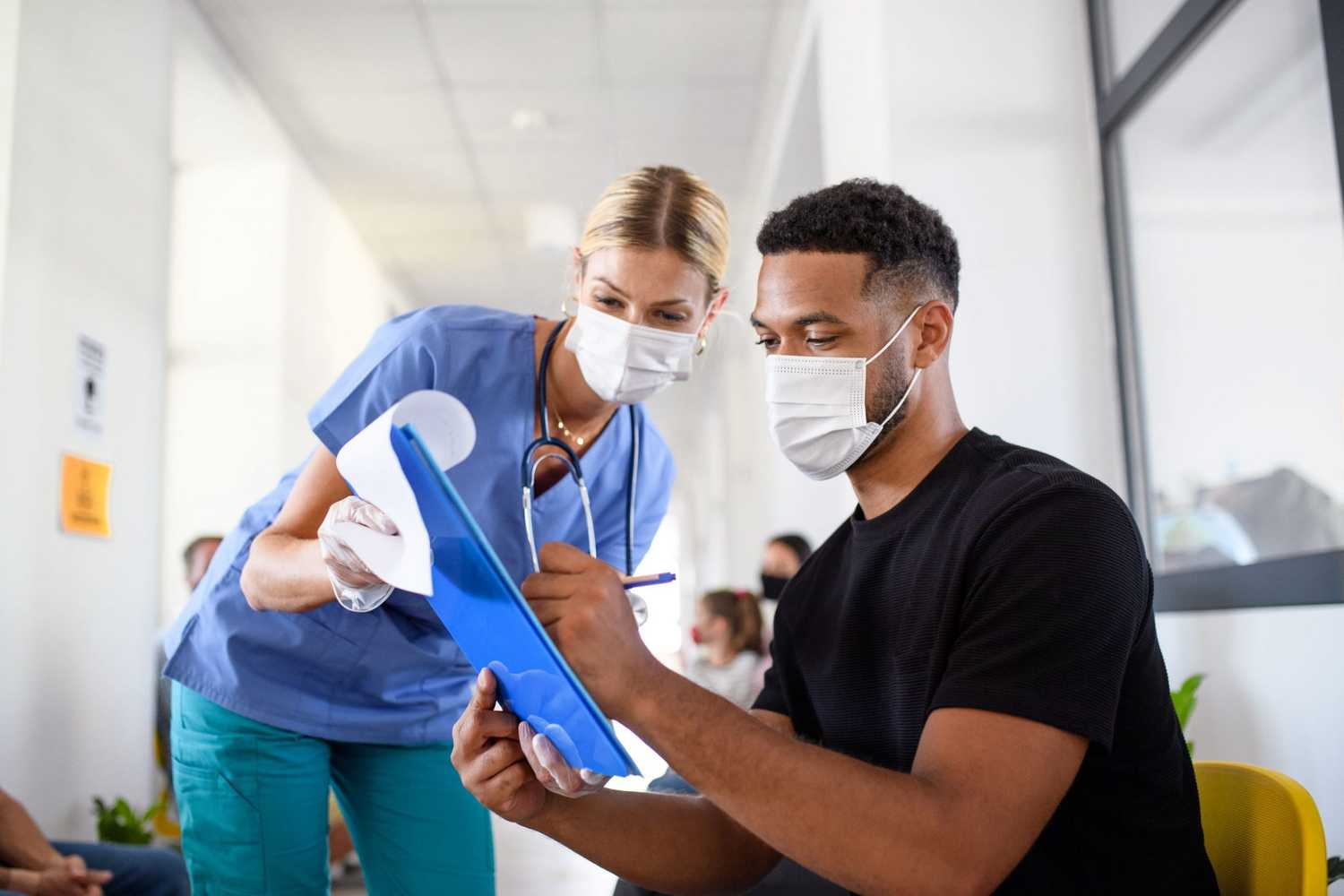 A doctor wearing a mask, helping a patient fill out a form, who is also wearing a mask