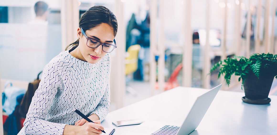 A woman in an office setting, sitting at a desk and writing something down