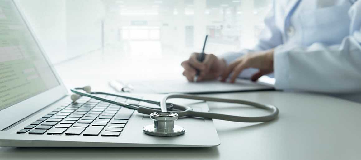 A doctor writing notes from a telehealth appointment next to his computer