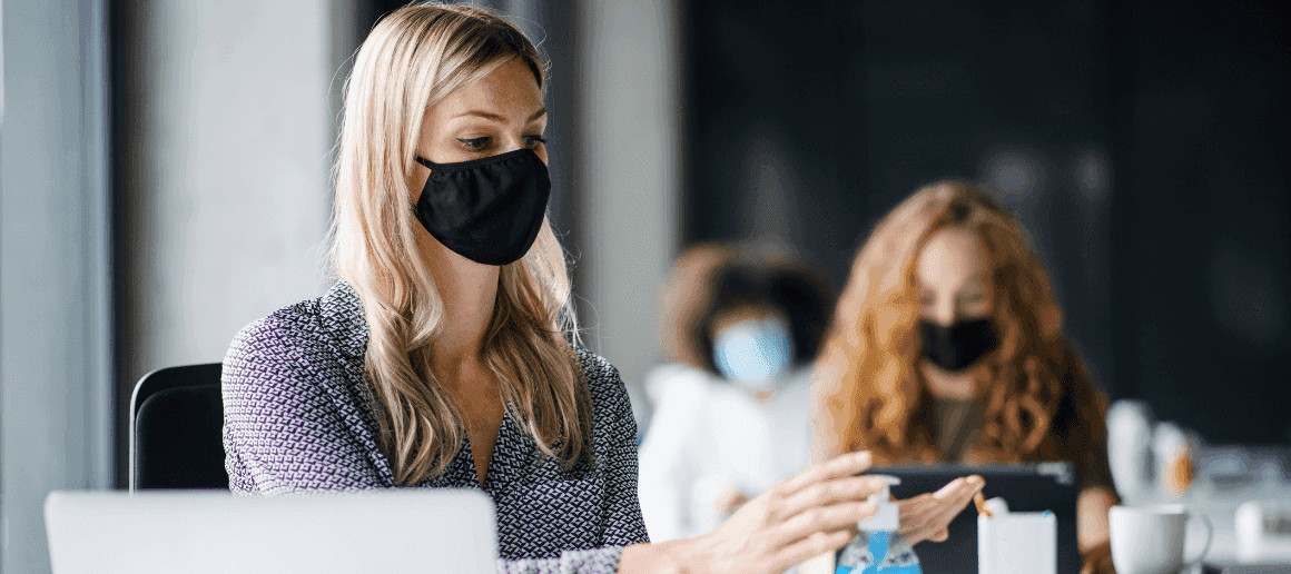 Three women sitting in an office wearing masks. The woman in the front is putting on hand sanitizer.