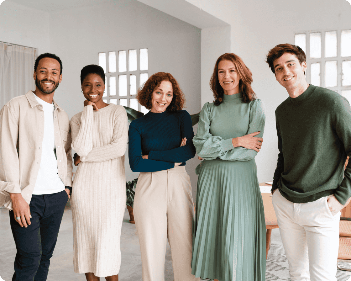 Image of five people standing in a line in various poses wearing cream colors, blues, and green shades of clothing smiling to camera in a white office setting